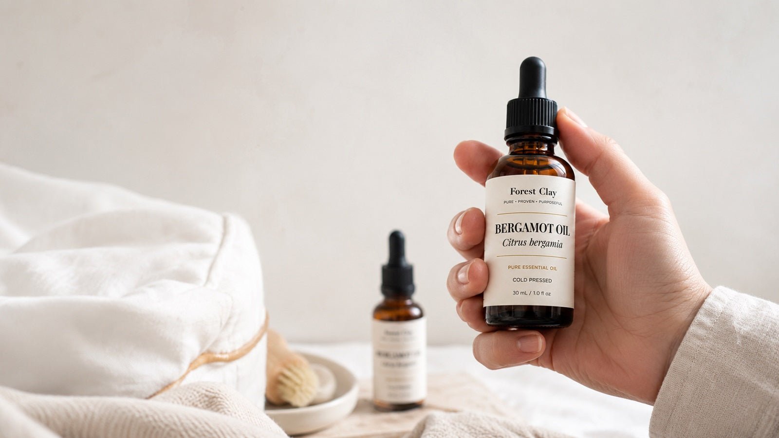 A Forest Clay bergamot essential oil bottle held beside soft natural textiles.